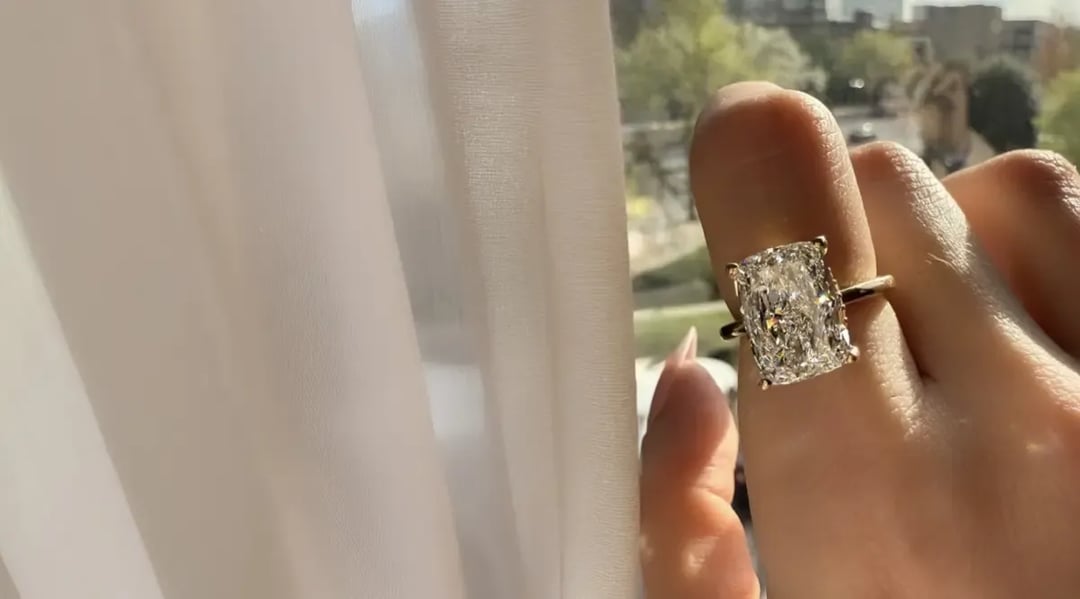 a ladies index finger is being shown against a window next to a white curtain. it is a a 4 prong elongated cushion diamond set in a yellow gold solitare engagement ring.